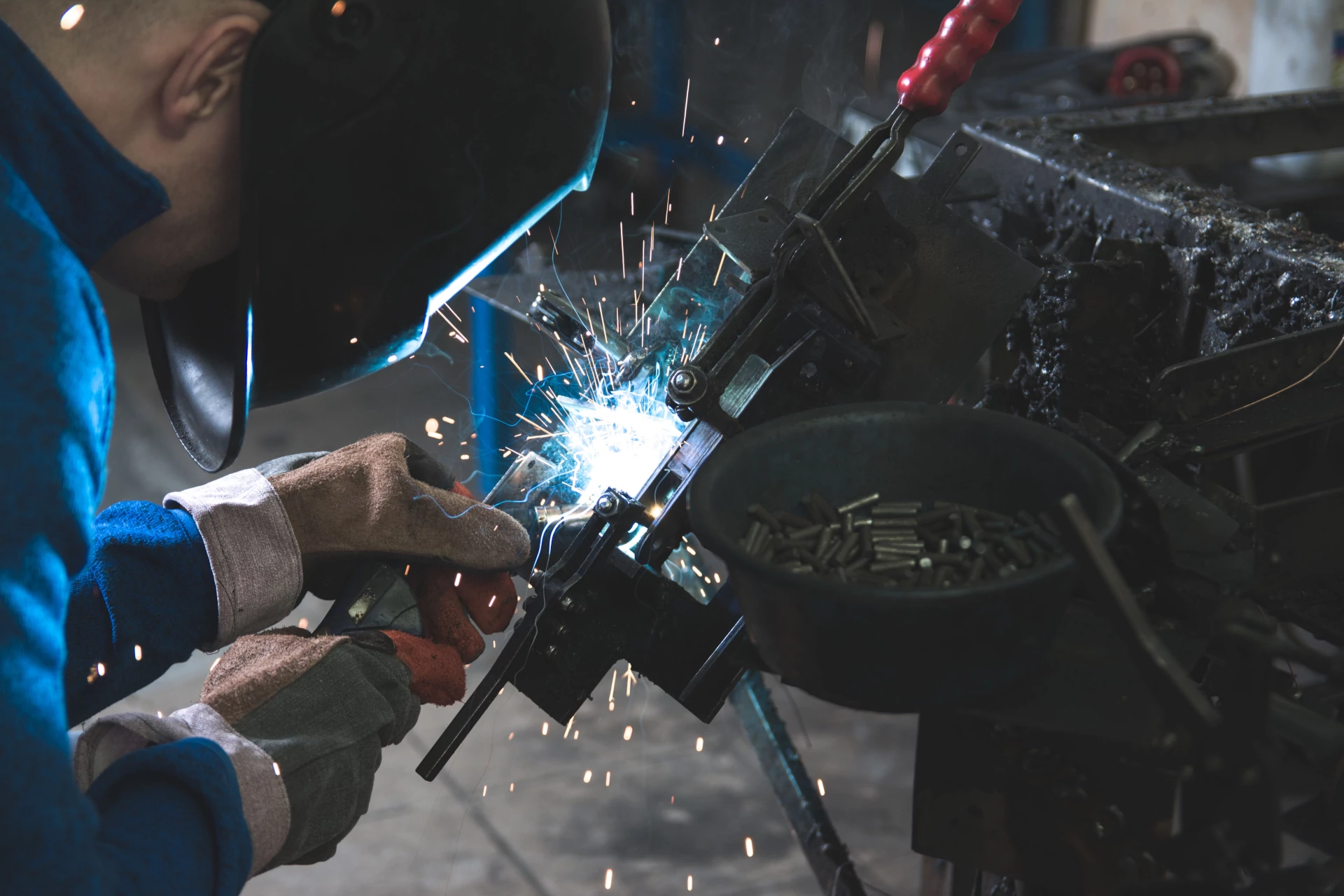 En esta imagen aparece un trabajador de la industria pesada soldando una pieza usando para ello diferentes equipos de protección individual tales como una careta de soldadura, unos guantes y un peto de trabajo de cuerpo completo